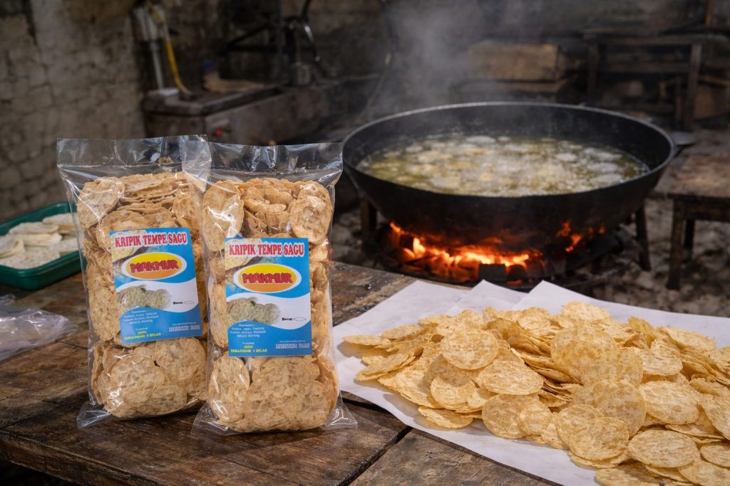 Keripik tempe sagu "Makmur" yang telah dikemas dalam plastik terlihat di atas meja, dengan latar proses produksi di dapur sederhana. (Foto: Dian Wahyu)