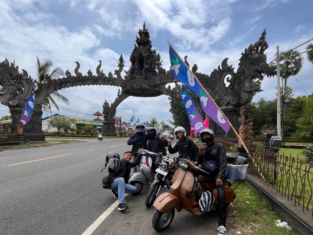 Potret pemuda pecinta Vespa berpose di bawah gapura Gilimanuk dengan latar langit cerah, menandai momen kebersamaan mereka saat touring yang penuh semangat. (Foto: Dian Wahyu)